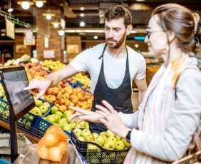 Handsome worker in uniform helping young woman customer to weigh apples on the scales in the supermarket