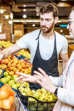 Handsome worker in uniform helping young woman customer to weigh apples on the scales in the supermarket